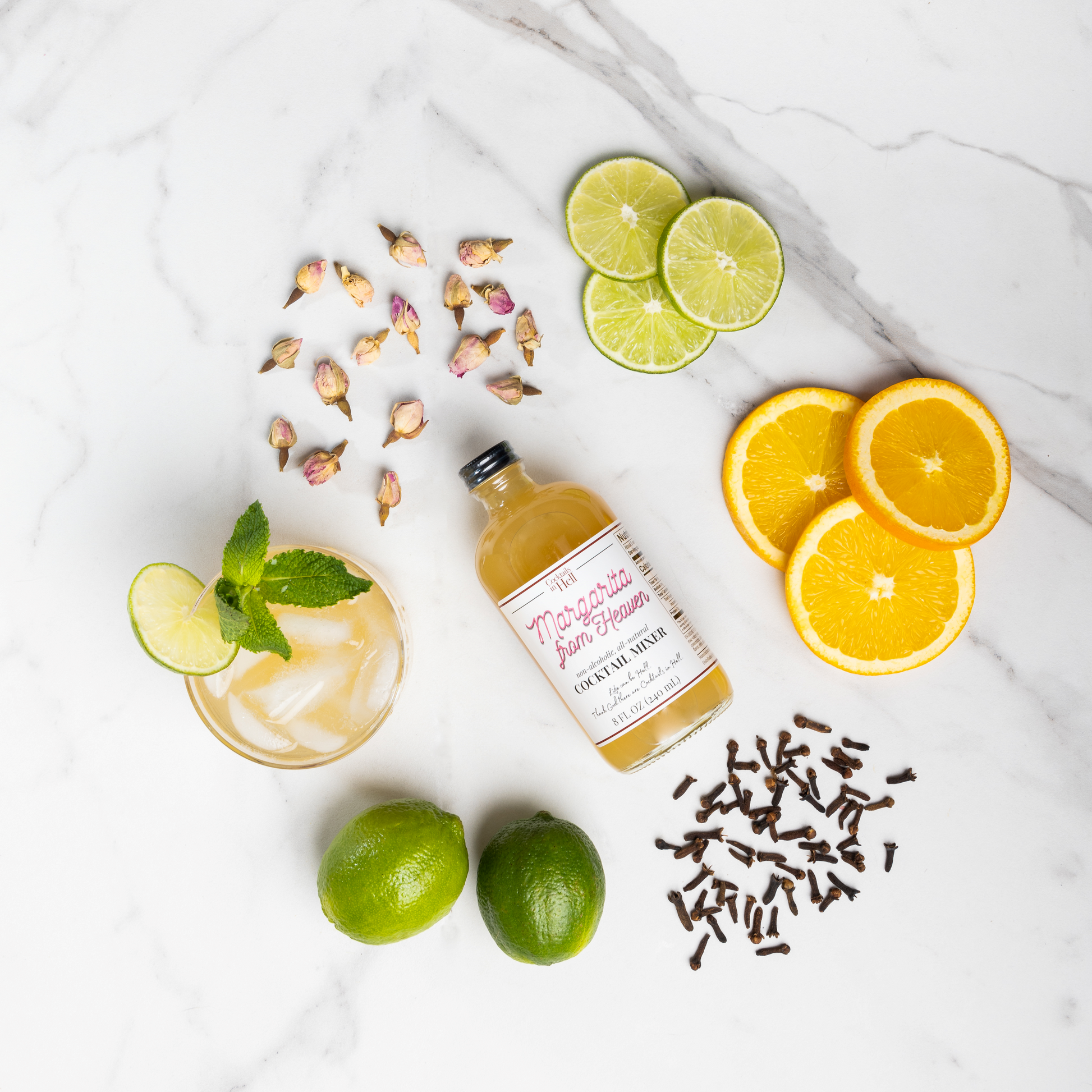 Glass bottle of Cocktails in Hell Margarita from heaven cocktail mixer next to lime slices, orange slices, spices and a glass of cocktail mixer